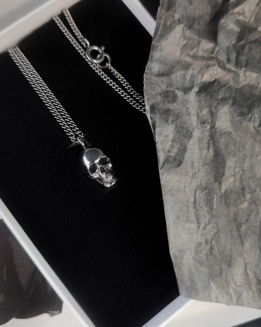 A silver skull pendant 16mm necklace on a chain, displayed inside a jewelry box next to crumpled gray paper.
