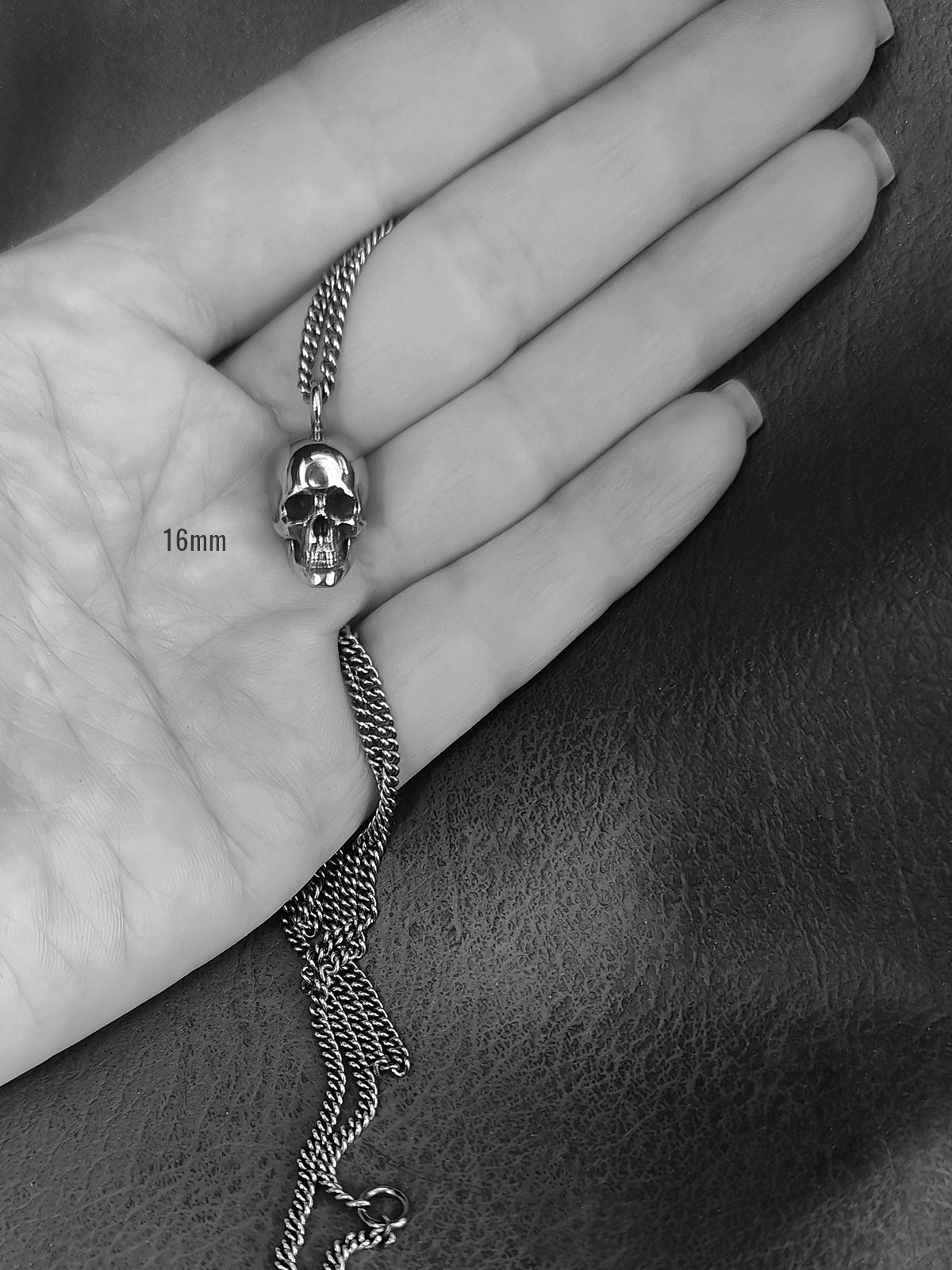 A silver skull pendant necklace on a chain, held in a person's hand. The image is in grayscale and features a '16mm' size overlay near the pendant.