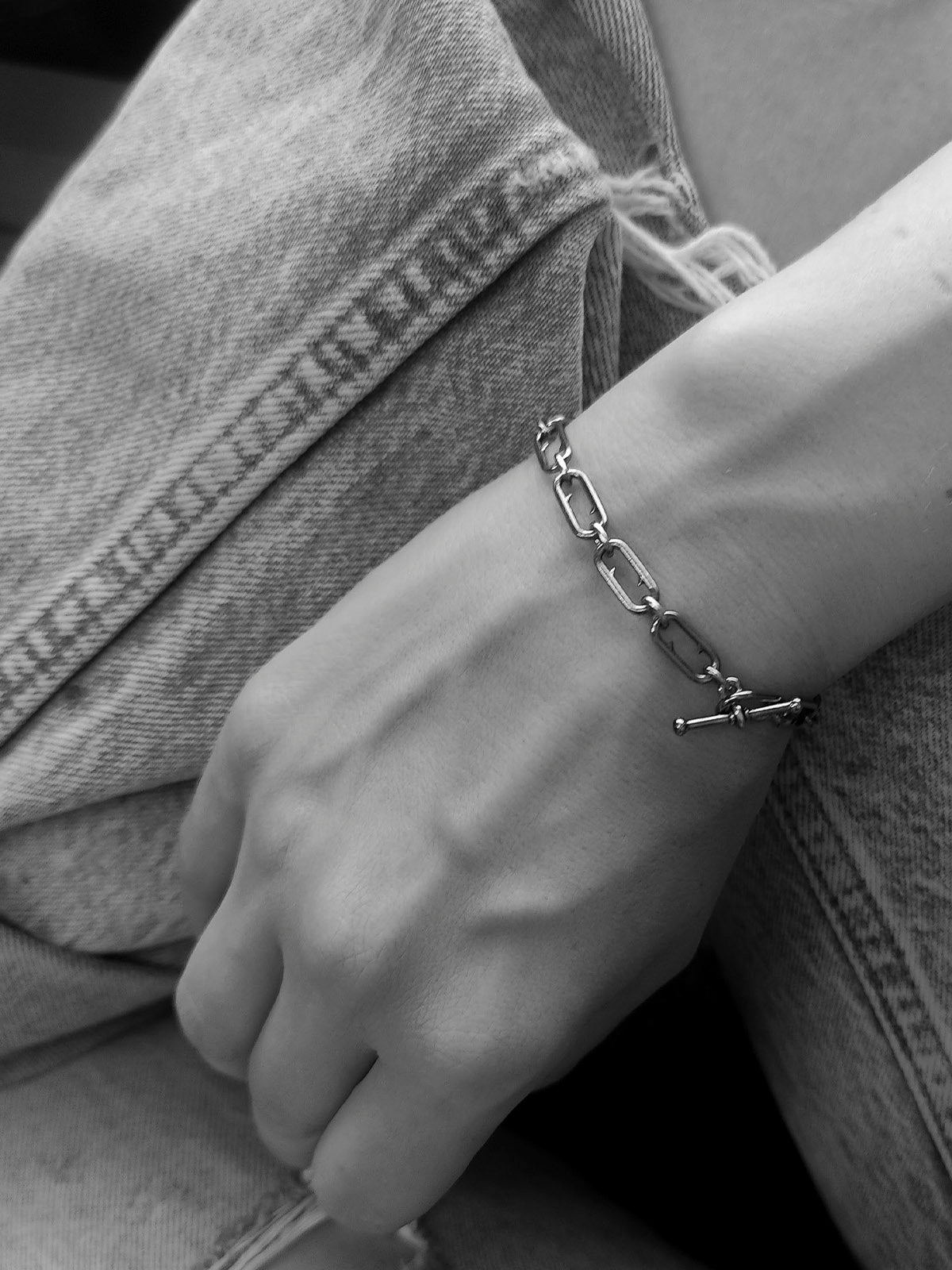 A close-up black and white photo of a person's wrist wearing a silver chain link bracelet with a toggle clasp, resting against denim fabric.