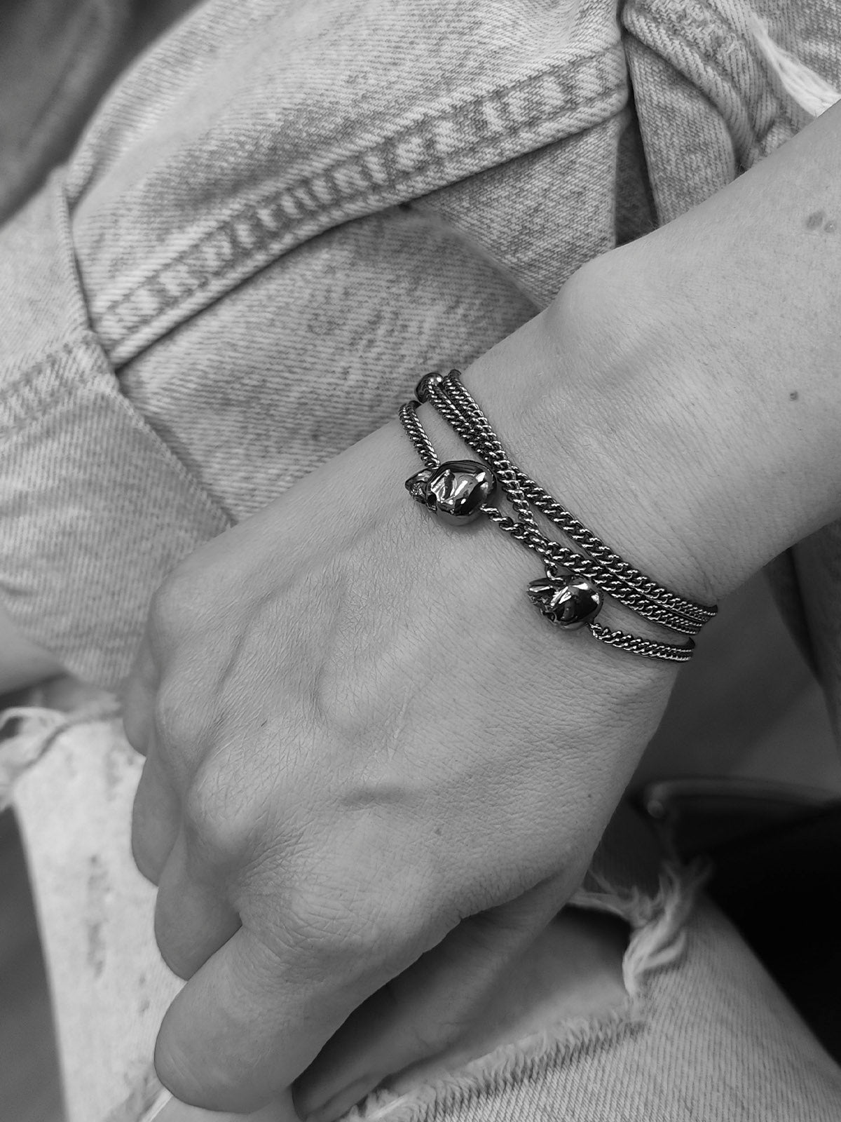 A close-up of a person's wrist wearing three layered metal bracelets, including 3 skull charms and textured chain straps, shown in black and white with denim clothing in the background.